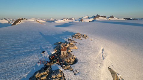 Eine kleine Berghütte steht auf einer Felskuppe mitten in einer weiten, schneebedeckten Gletscherlandschaft, umgeben von Bergen im goldenen Licht der tiefstehenden Sonne.