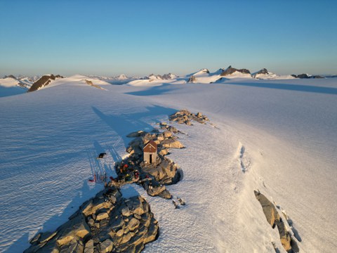 Eine kleine Berghütte steht auf einer Felskuppe mitten in einer weiten, schneebedeckten Gletscherlandschaft, umgeben von Bergen im goldenen Licht der tiefstehenden Sonne.