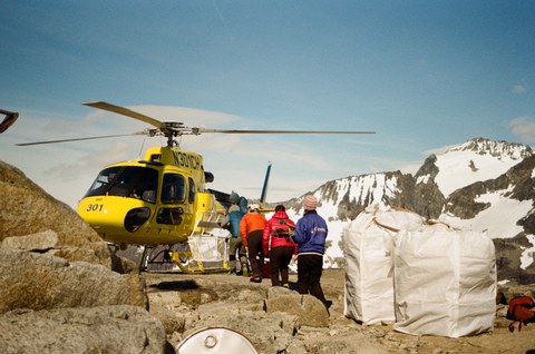 Eine Gruppe von Menschen nähert sich einem gelben Hubschrauber, der auf felsigem Untergrund in einer schneebedeckten Berglandschaft gelandet ist.