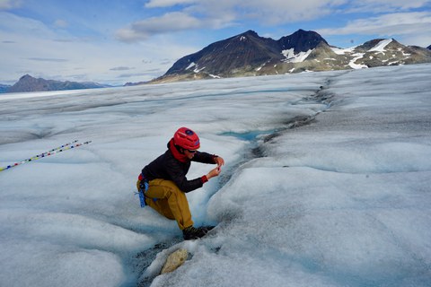 Eine Forscherin oder ein Forscher kniet auf einem Gletscher und entnimmt eine Probe aus einer kleinen Schmelzwasserrinne. Im Hintergrund sind Berge und Schneefelder zu sehen.
