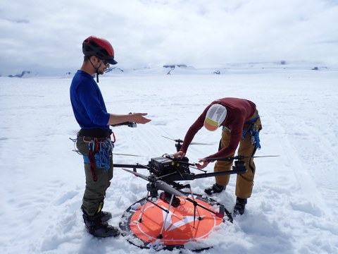 Zwei Forschende bereiten auf einem verschneiten Gletscher eine große Drohne für eine LiDAR-Vermessung vor. Beide tragen Helme, Klettergurte und Winterausrüstung.