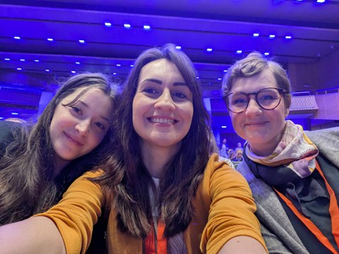 Selfie von drei Frauen in einem Saal vor der Bühne und lächeln in die Kamera