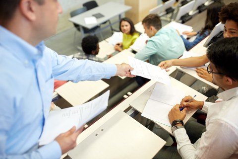 Studierende sitzen in einem Hörsaal. Ein Dozent teilt ihnen Arbeitsblätter aus.