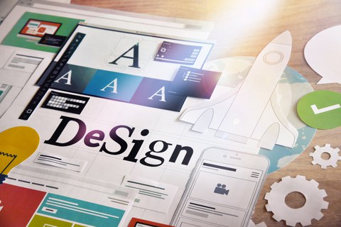 A collage of design elements on a table. The word "Design" is prominently displayed in the foreground, surrounded by typographic samples, graphics, illustrations, and icons such as a rocket, a gear, a light bulb, and a smartphone. 