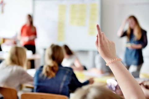 Unscharf erkennt man Menschen in einem Klassenzimmer. Eine Person meldet sich.