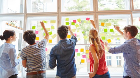 5 Personen stehen am Fenster und befestigen Klebzettel auf den Scheiben - Methode Brainstorming.