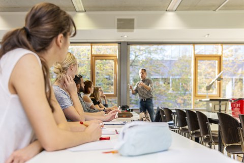 Studierende sitzen in einem Seminarraum und hören einem Mann zu.