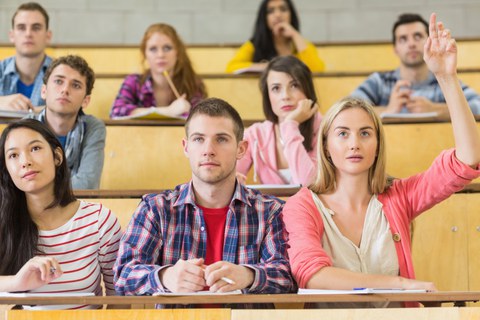 Das Foto zeigt mehrere Studierende während einer Veranstaltung in einem Hörsaal. Eine Studentin hebt gerade ihre Hand.