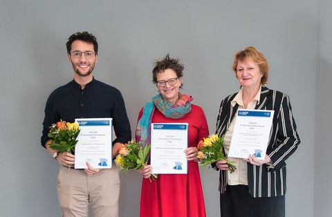  Foto: Ein Mann und 2 Frauen stehen nebeneinander und haben eine Urkunde und einen Blumenstrauß in der Hand