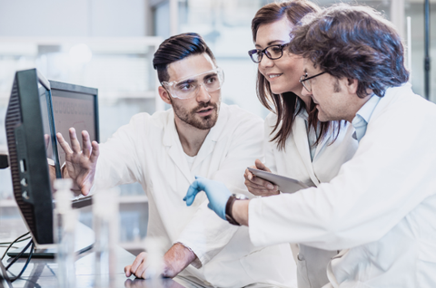 A team of three scientists in white lab coats is working together at a computer in a laboratory. The man on the left is pointing at the screen, while the woman in the middle is looking at a tablet, and the man on the right is listening attentively. They are all wearing safety goggles, and there are lab utensils on the table in front of them.