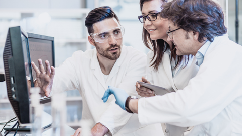 A team of three scientists in white lab coats is working together at a computer in a laboratory. The man on the left is pointing at the screen, while the woman in the middle is looking at a tablet, and the man on the right is listening attentively. They are all wearing safety goggles, and there are lab utensils on the table in front of them.