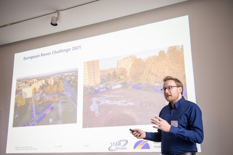 A man is giving a presentation. His PowerPoint can be seen the background.