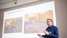 A man is giving a presentation. His PowerPoint can be seen the background.