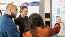 3 students are standing in front of a scientific poster. One student is pointing at a diagramm on the poster.