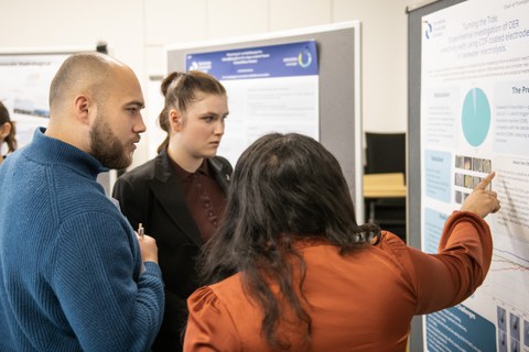3 students are standing in front of a scientific poster. One student is pointing at a diagramm on the poster.