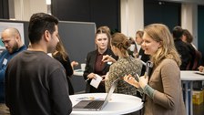 Mehrere Personsn stehen verteilt an verschiedenen Stehtischen und unterhalten sich.