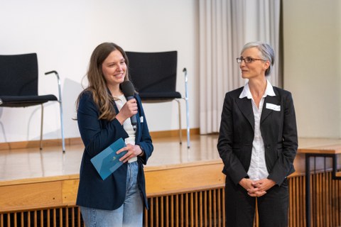 Eine Frau spricht in ein Mikrofon und hält in der anderen Hand Moderationskarten. Rechts neben ihr steht noch eine Frau, die den Blick auf die Sprecherin gerichtet hat.