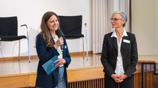 Eine Frau spricht in ein Mikrofon und hält in der anderen Hand Moderationskarten. Rechts neben ihr steht noch eine Frau, die den Blick auf die Sprecherin gerichtet hat.