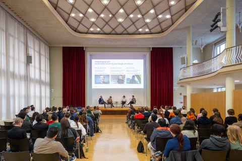 Links und rechts sitzt das Publikum. Vorne auf der Bühne sitzen 4 Leute und diskutieren bei einem Podiumsgespräch. 