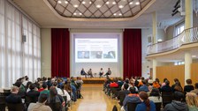 Links und rechts sitzt das Publikum. Vorne auf der Bühne sitzen 4 Leute und diskutieren bei einem Podiumsgespräch. 