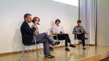 Seitlicher Blick auf die Bühne und das Podiumsgespräch. Der Mann, der ganz links sitzt, spricht in das Mikrofon.
