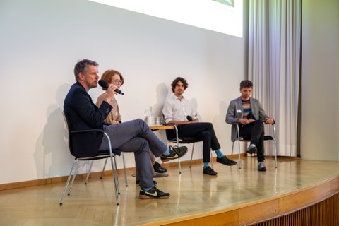 Seitlicher Blick auf die Bühne und das Podiumsgespräch. Der Mann, der ganz links sitzt, spricht in das Mikrofon.