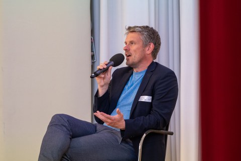 Ein Mann sitz zurück gelehnt in einem Stuhl und spricht in ein Mikrofon.
