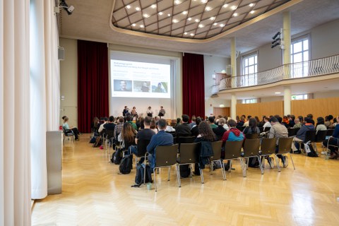 Das Bild zeigt das Publikum von hinen. Vorne ist eine Präsention and die Wand über der Bühne geworfen.