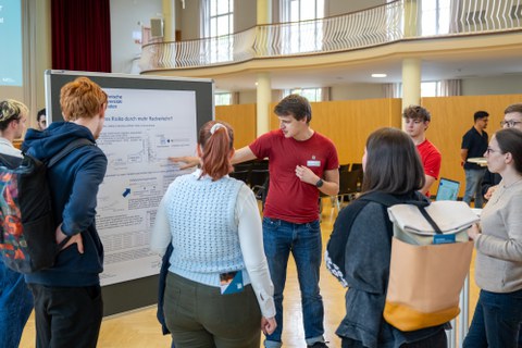 Mehrere Menschen stehen vor einer Pinnwand mit Poster, ein Student erklärt etwas und zeigt auf das Poster.