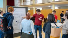 Mehrere Menschen stehen vor einer Pinnwand mit Poster, ein Student erklärt etwas und zeigt auf das Poster.