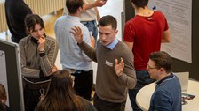 Das Bild zeigt mehrere Menschen, die zusammenstehnen und sich unterhalten. Die Person in der Mitte gestikuliert und macht seine Arme auf.