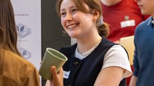 Zwei Personen sprechen miteinander. Die Person, die von hinten zu sehen ist hält einen Becher in ihrer rechten Hand.