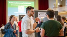 Ein Student und eine Studentin unterhalten sich. Im Hintergrund sind mehr Menschen zu sehen, die sich die Poster anschauen.
