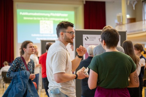 Ein Student und eine Studentin unterhalten sich. Im Hintergrund sind mehr Menschen zu sehen, die sich die Poster anschauen.