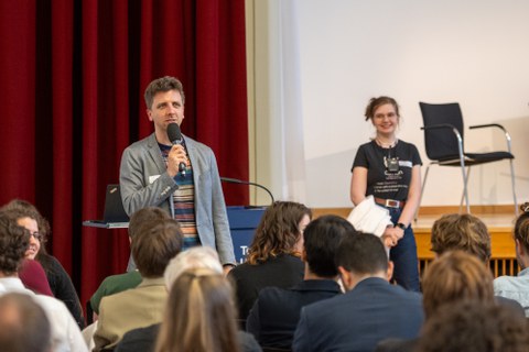 Unten ist das Publikum von hinten zu sehen. Vorne steht der Moderator und neben ihm ein Studentin.