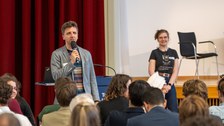 Unten ist das Publikum von hinten zu sehen. Vorne steht der Moderator und neben ihm ein Studentin.