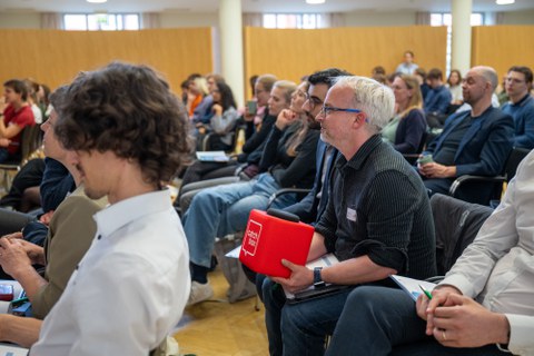 Ein Mann im Publikum hält eine rote Mikrofonbox und stellt eine Frage.