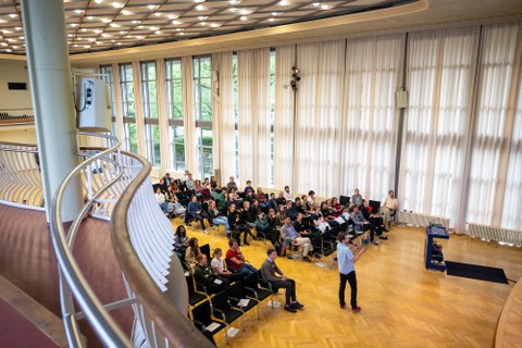Das Bild zeigt den Raum von der Galerie von oben aus. Unten sieht man das Publikum sitzen und einen Studenten der vorne steht und einen Vortrag hält.