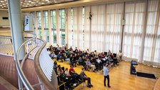 Das Bild zeigt den Raum von der Galerie von oben aus. Unten sieht man das Publikum sitzen und einen Studenten der vorne steht und einen Vortrag hält.