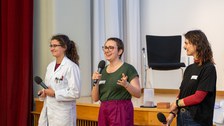 Drei Studentinnen stehen vorne. Die Person links trägt einen Laborkittel. Die Person in der Mitte spricht in ein Mikrofon.
