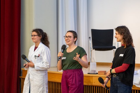 Drei Studentinnen stehen vorne. Die Person links trägt einen Laborkittel. Die Person in der Mitte spricht in ein Mikrofon.