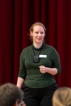 Eine Studentin in grünem Pulli hält einen Vortrag. Sie steht vor einem roten Vorhang.