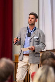 Ein Student hält einen Vortrag. Das Publikum ist unten im Bild verschwommen zu sehen.