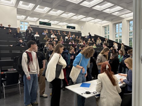 Students gather in a lecture hall at TU Dresden. In the foreground, several people register at a table with two staff members, while many other students take their seats in the background.