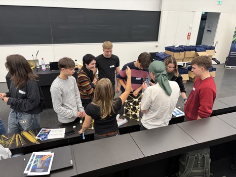 Students work in groups on a building project made of spaghetti and marshmallows in a lecture hall.