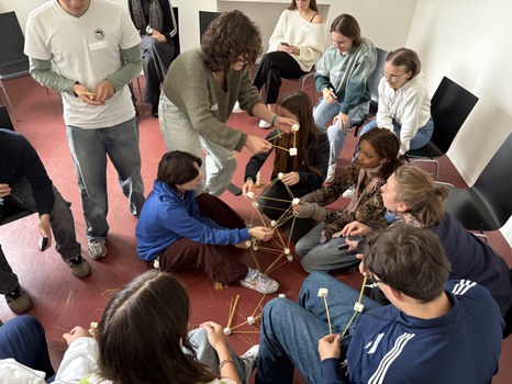 Students work together to build a tower out of spaghetti and marshmallows. Some are sitting, others are standing and actively helping to build.