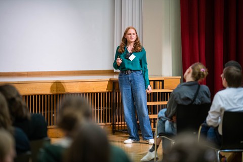 Eine junge Frau steht auf einer Bühne in einem Hörsaal und hält eine Präsentation vor sitzenden Zuhörer:innen. Sie spricht frei und blickt ins Publikum.