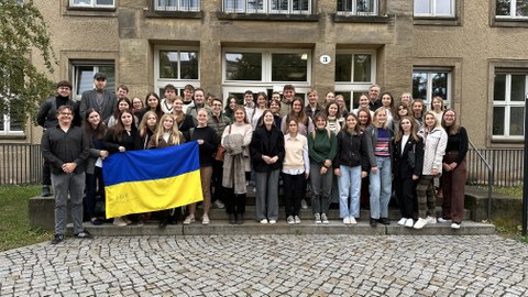 Student:innen aus der Ukraine und Deutschland stehen vor einem brauen Universitätsgebäude. Drei Studenten halten eine Ukraine-Flagge hoch