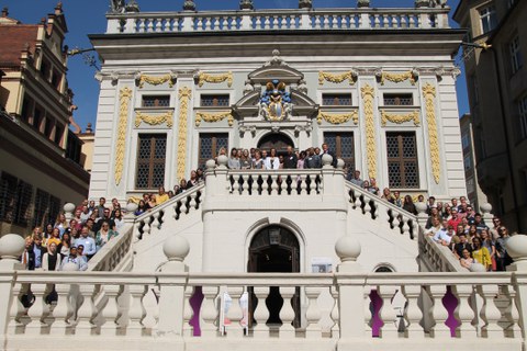 Frontales Foto eines barocken Gebäudes. Auf der Treppe zum Eingang befinden sich etwa einhundert Studierende