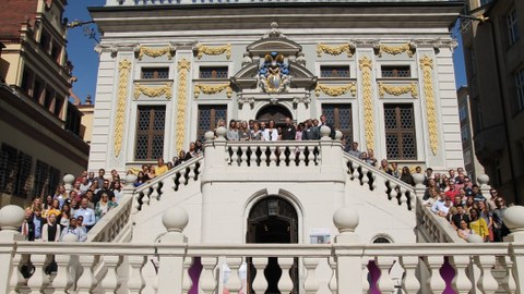 Frontales Foto eines barocken Gebäudes. Auf der Treppe zum Eingang befinden sich etwa einhundert Studierende